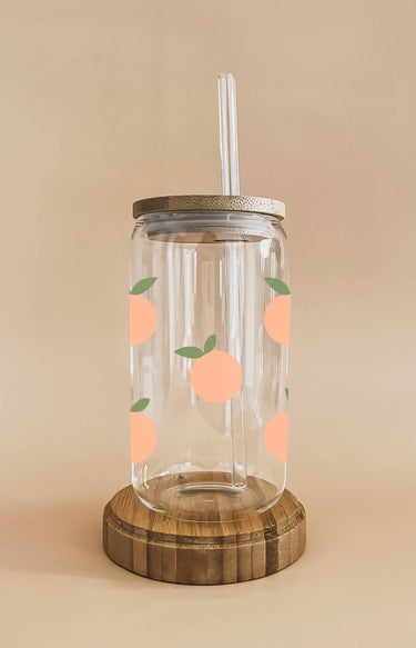 A clear glass cup with a bamboo lid and straw is decorated with light orange peach decals with green leaves and rests on a round wooden coaster.